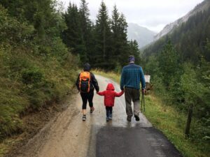 two adults and one child hold hands while walking through the rain on a hike, away from the camera