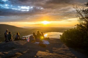 a group of people sit overlooking a lake and enjoy a picnic dinner as the sun sets. Image for a blog post on the importance of community.